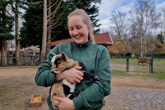 Caroline Otto steht in einem Gehege im Tierpark und lächelt auf eine kleine Ziege in ihren Armen herab.