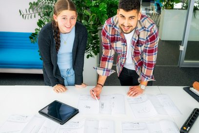 Ein junger Mann und eine junge Frau sprechen über Konstruktionszeichnungen, die vor ihnen auf dem Tisch liegen.