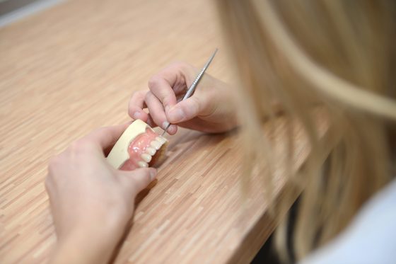	Eine Frau hält Skalpell mit sehr dünner Klinge ab ein künstliches Gebiss über einen Tisch.