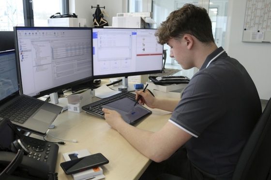 Ein junger Mann sitzt mit einem Tablet vor einem Computer und arbeitet.