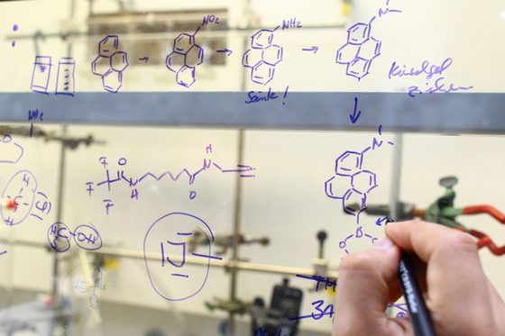 Im Labor sind chemische Formeln und Elemente auf eine Glaswand geschrieben. Eine Hand hält einen blauen Stift.