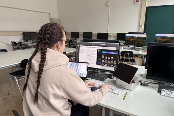 Rückansicht einer jungen Frau am Arbeitsplatz, die vor mehreren Bildschirmen sitzt und arbeitet. 