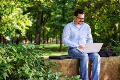 Ein junger Mann sitzt mit einem Laptop auf einer Mauer im Park. Foto: Julien Fertl