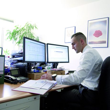 Ein junger Mann sitzt an einem Schreibtisch vor einem Computer und hat eine Mappe vor sich aufgeschlagen.
