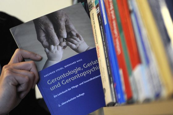 Eine Hand kommt vom linken Bildrand in das Foto und nimmt ein Buch über Gerontologie aus einem Bücheregal.