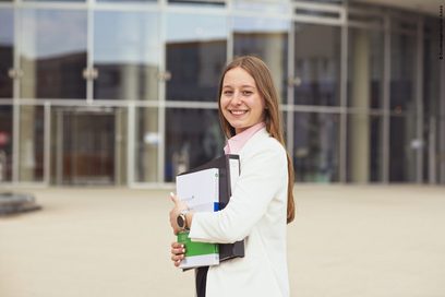Porträt einer jungen Frau, die Schulungsunterlagen trägt, vor einem Bürogebäude.