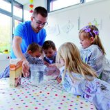 Ein junger Mann mit Brille, bekleidet mit einem blauen T-Shirt leitet zwei Mädchen und zwei Jungen, an einem Tisch mit bunt gepunkteter Tischdecke, bei der Herstellung eines Knetteiges an. Die Kinder haben die Hände in einer Mehlschüssel, der junge Mann greift an einen Messbecher mit Wasser.