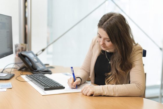 Eine Frau sitzt an einem Schreibtisch und notiert etwas auf einem Zettel.