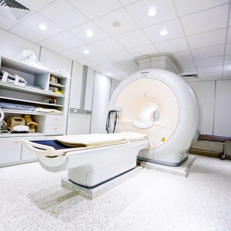 In einem Behandlungszimmer steht ein großes weißes MRT-Gerät. An der Seite sind Regale und Schränke angebracht, die weiteres medizinisches Equipment beherbergen.