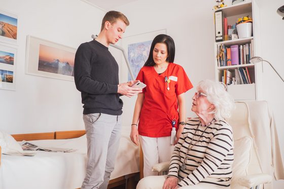 Ein junger Mann zeigt einer Pflegerin und einer älteren Dame etwas auf einem Smartphone.