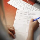 Eine Person sitzt vor einem Tisch, auf dem eine Computertastatur liegt. Die langen Haare der Person sind am linken Bildrand zu sehen. Sie schreibt mit einem Stift auf ein Blatt Papier.