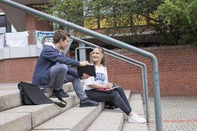 Ein Schüler und eine Schülerin sitzen auf einer Treppe vor einer Schule.