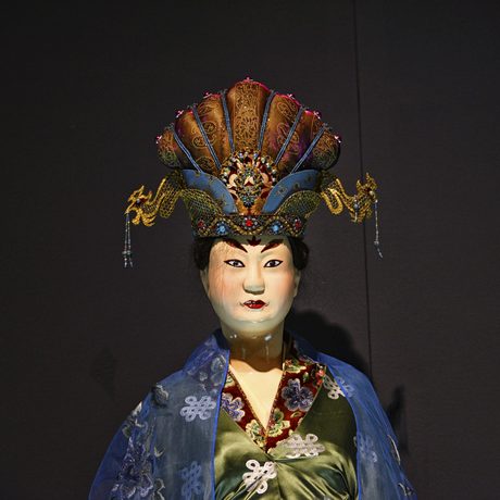 Eine chinesische Figur aus Terrakotta in traditionellem Gewand steht vor einem dunklen Hintergrund.