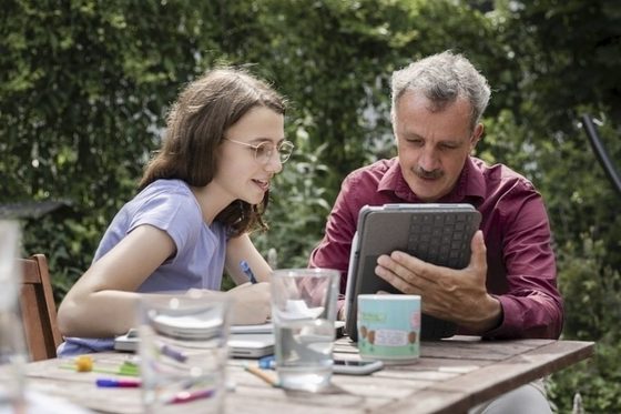 Ein Vater und seine Tochter sitzen an einem Tisch und blicken auf ein Tablet.