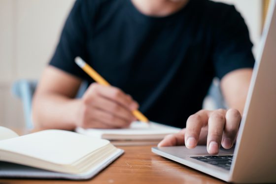Ein Mann betätigt die Pfeiltaste eines Laptops und schreibt etwas auf Papier.