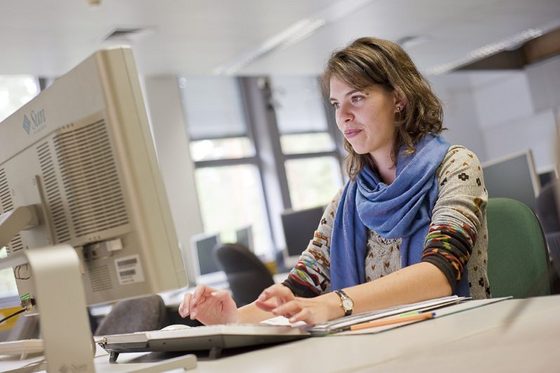 Auf dem Foto ist eine junge Frau zu sehen, die an einem Rechner sitzt. Sie hat helle, lockige Haare, trägt ein bunt gemustertes Oberteil und ein blaues Tuch um den Hals. Sie sitzt in einem Computerraum einer Hochschule.