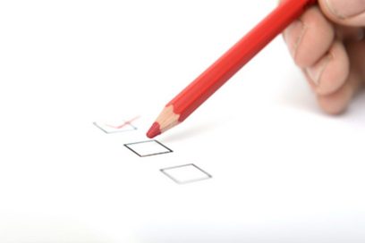 Eine Checkliste mit drei schwarzen quadratischen Kästchen von denen das obere mit einem roten Buntstift angehakt wurde. Die Hand einer Person hält den roten Buntstift und zeigt auf das mittlere Kästchen.