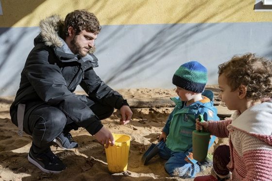 Ein junger Mann spielt mit zwei Kindern in einem Sandkasten.