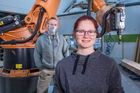 Eine junge Frau und ein Mann stehen vor einem Produktionsroboter.