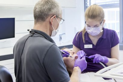 Eine junge Frau mit Schutzbrille und Mundschutz, bekleidet mit einem violetten Poloshirt, assistiert einem Zahnarzt