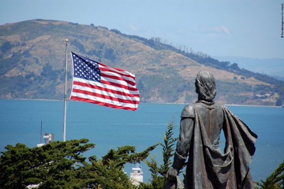 Im Vordergrund die Statue von Christoph Kolumbus von hinten mit Blick auf die Bucht vor ihm. Links weht die Flagge der Vereinigten Staaten.