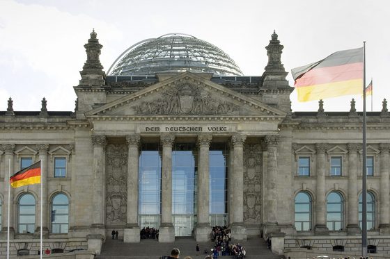 Aufnahme des Reichstagsgebäudes mit Glaskuppel in Berlin. Auf und vor dem Gebäude stehen mehrere Deuschlandflaggen und eine Europaflagge.