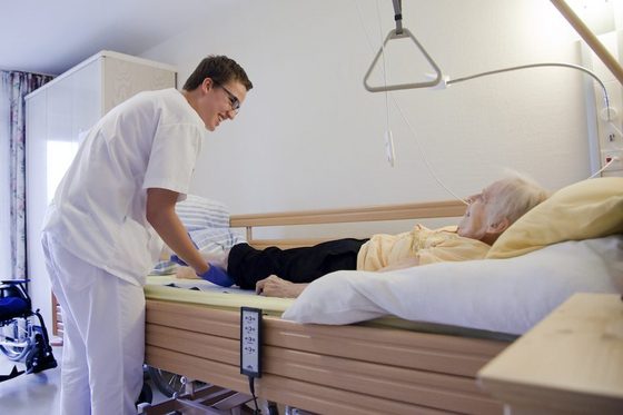 Ein junger Mann in weißer Pflegerkleidung steht am Pflegebett einer liegenden älteren Frau.