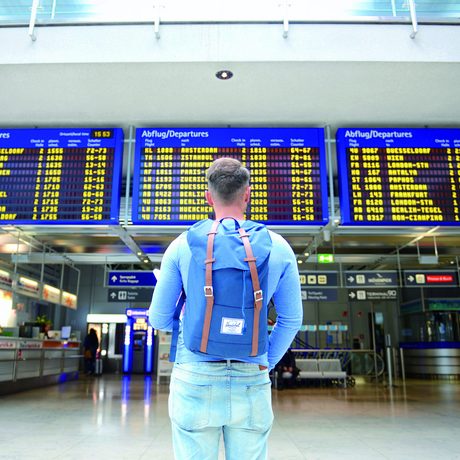 Aufnahme eines jungen Mannes von hinten, er hat kurze Haare, ist bekleidet mit einer blauen Jeans, einem blauen Langarmshirt, hat einen blauen Rucksack auf dem Rücken und steht vor den Anzeigetafeln einer Abflughalle in einem Flughafen.