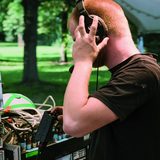 Ein junger Mann mit Kopfhörer steht im Freien auf einer Wiese vor einer Musikanlage, und prüft einen Anschluss.