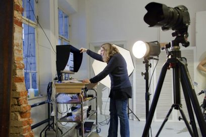 Eine Frau sieht in einen großen Bildschirm auf einem Rolltisch, während im Vordergrund eine Kamera steht.