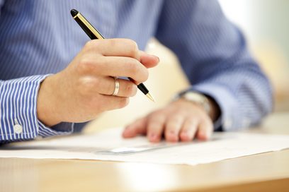Eine Männerhand hält einen Kugelschreiber über ein Lineal auf einem Dokument, das auf einem Schreibtisch liegt.  (Foto: Swen Reichhold | Bundesagentur für Arbeit)