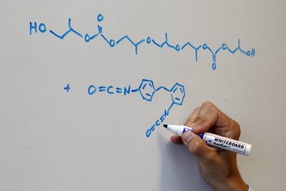 An einem Whiteboard sind chemische Formeln als Kettenverbindung aufgeschrieben. Eine Hand hält einen blauen Whitboardmarker an eine der Formeln.