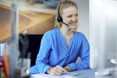 Eine junge Frau mit Headset sitzt an ihrem Schreibtisch und lächelt.