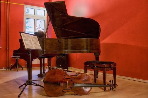 Zu sehen ist ein rot gestrichener Raum mit einem Flügel, davor stehen ein Notenpult und ein Klavierhocker, im Vordergrund liegt ein Cello.
