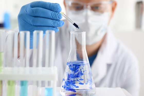 Chemiker mit Schutzbrille, Mundschutz und Gummihandschuhen tropft mit einer Pipette eine blaue Flüssigkeit in einen Erlenmeyerkolben, der mit einer klaren Flüssigkeit gefüllt ist.