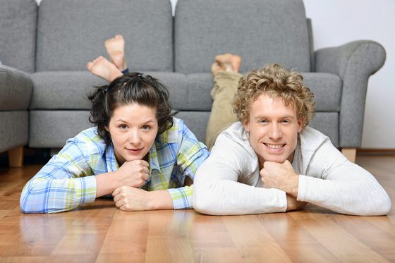 Eine junge Frau und ein junger Mann liegen bäuchlings und mit nackten Füßen auf einem Holzfußboden vor einem grauen Sofa. Beide lächeln in die Kamera, wobei sie ihren Kopf auf ihre zu Fäusten geballten Hände stützen.