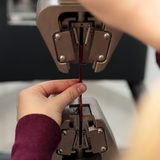 Zwei Hände führen ein längliches Kunststoffprobeteil durch eine Festigkeitsprüfmaschine