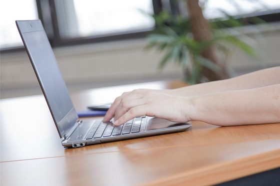 Ein Notebook liegt auf einem holzfarbenen Tisch. Auf der Tastatur liegen zwei Hände. Im Hintergrund ist eine Pflanze erkennbar.