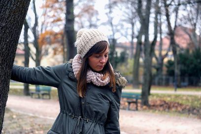 Zu sehen ist eine junge Frau in einem Park, gekleidet mit einem grünen Parka, Wollschal und Wollmütze. Sie hält sich mit einer Hand an einem Baumstamm fest und blickt betrübt zu Boden.