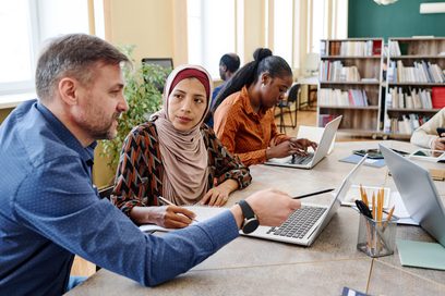 Ein Lehrer erklärt einer erwachsenen muslimischen Schülerin mit Hidschab etwas an dem Laptop vor den die beiden sitzen. 