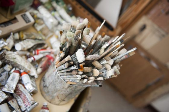 Aufnahme einer Dose aus der Vogelperspektive, in der sehr viele gebrauchte Pinsel stehen. Um die Dose herum liegen auf einem Tisch sehr viele Farbtuben.