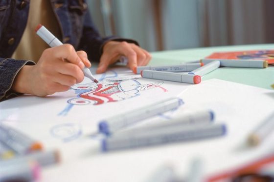 Eine Frau sitzt am Schreibtisch. Vor ihr liegt ein Blatt Papier mit einer Motorradzeichnung. Auf der Zeichnung und auf dem Schreibtisch sind mehrere Textmarker in verschiedenen Farben zu sehen. Die Frau hält einen roten Stift in der Hand.