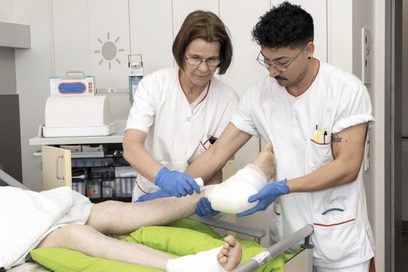 Ein junger Mann und eine Frau in medizinischer Arbeitskleidung behandeln einen Patienten mit Gipsfuß.