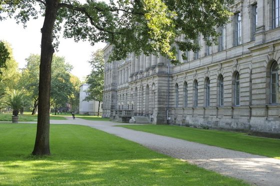 Ein großes altes Gebäude neben einem Park mit Wegen, Rasen und Laubbäumen. Im Hintergrund ist ein anderes helles Gebäude zu erkennen, ein Radfahrer und verschiedene Personen