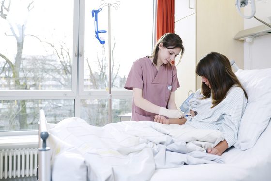 Eine junge Frau mit braunen Haaren sitzt in einem Krankenhausbett in einem lichtdurchfluteten Krankenzimmer vor einem Fenster. Neben ihr steht eine junge braunhaarige Krankenschwester in einem rosa Arbeitskittel und mit Handschuhe, die sie am rechten Arm für eine Infusion vorbereitet.