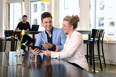 Ein junger Mann und einen junge Frau sitzen in einem Cafe an einem schwarzen Tisch mit gelber Tulpe in einer Glasblumenvase und schauen lachend auf ein Tablet. Auf dem Tisch stehen eine Kaffeetasse und eine Wasserglas. Im Hintergrund sitzt eine junger Mann mit eine Laptop an einem hohen Tisch.