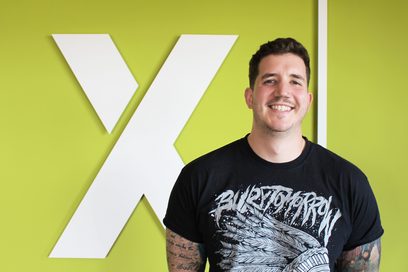 Ein junger Mann mit tattöwierten Armen steht vor einer hellgrünen Wand mit seinem Firmenlogo, einem großen weißen X, und lächelt.