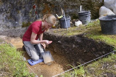 Eine Archäologin sitzt, mit einer Schaufel in der Hand, in einer Grube. (Foto: Frank Pieth)